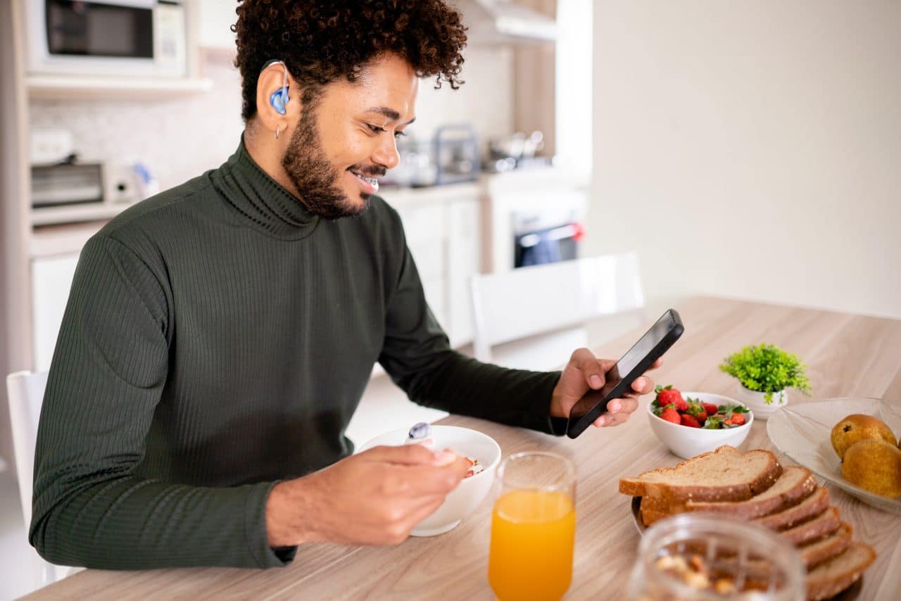 Young man with hearing aids checking his phone over breakfast.